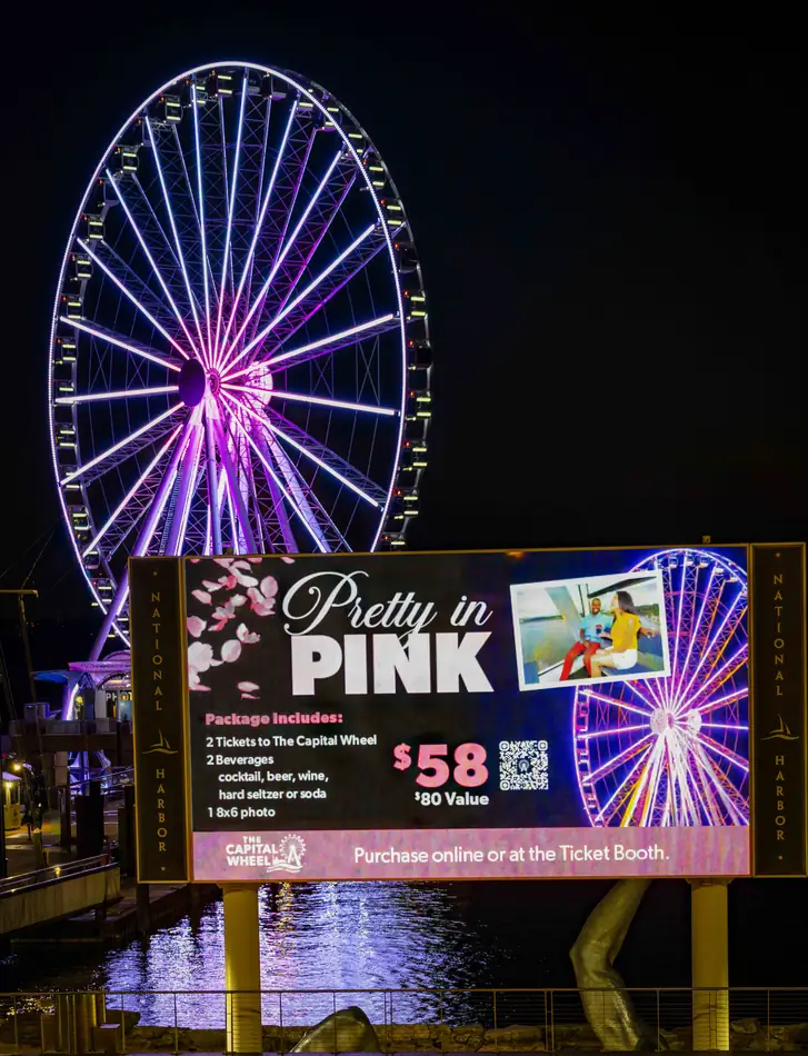 The Capital Wheel lit pink at night beside the Pretty in Pink campaign billboard featuring Tosh Persons photography at National Harbor